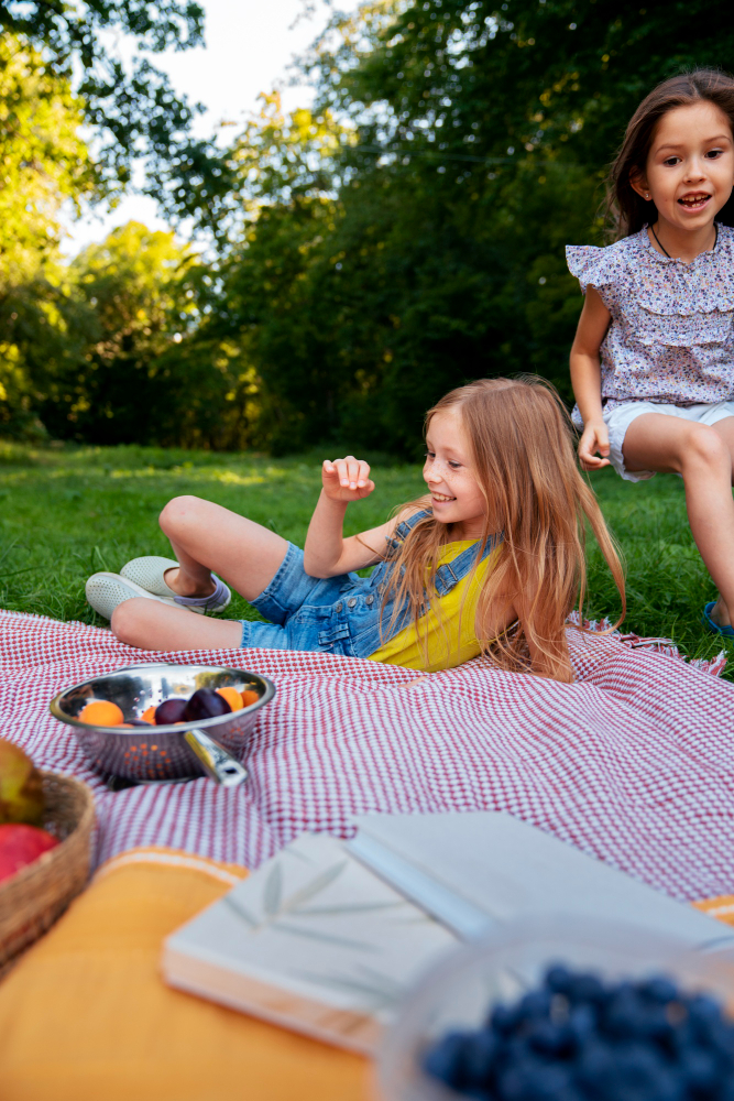 A young girl lounges happily on a checkered picnic blanket outdoors, with another girl running up beside her and fruit, a notebook, and berries in the foreground.